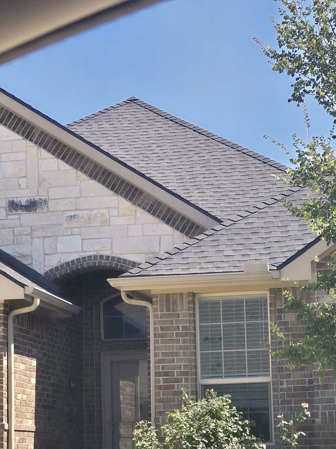 Gray shingles along the gable of a stone home, seen from below against a blue sky