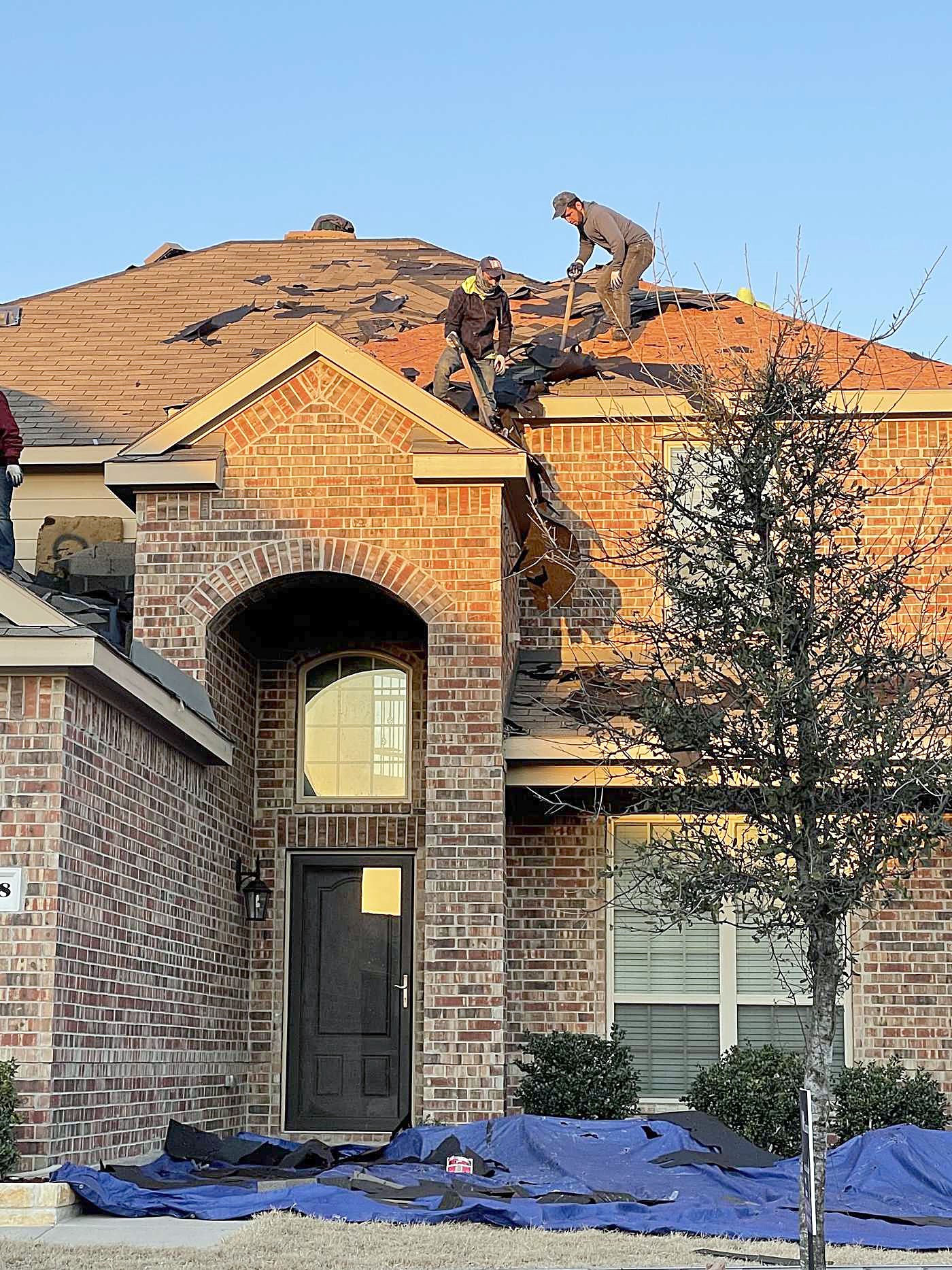 Roofers working on a brick home during a tear-off, with blue tarps protecting the yard
